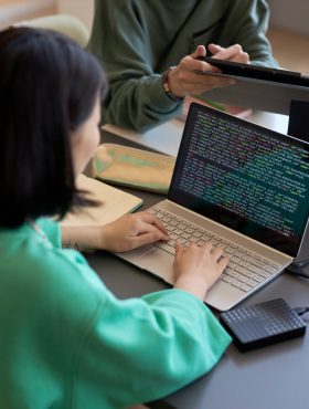 Young female student typing on laptop keyboard while decoding data on screen against two classmates or colleagues discussing presentation